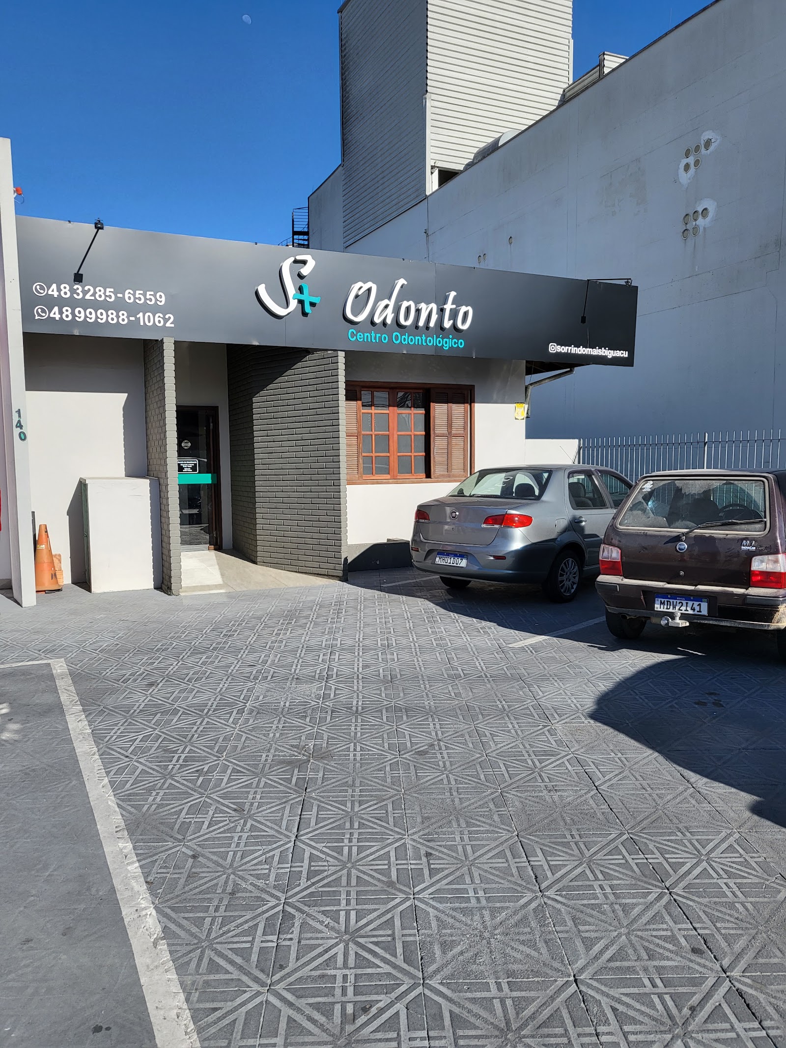 Fachada da clínica com letreiro escuro, entrada em tijolos cinza e vagas de estacionamento em frente