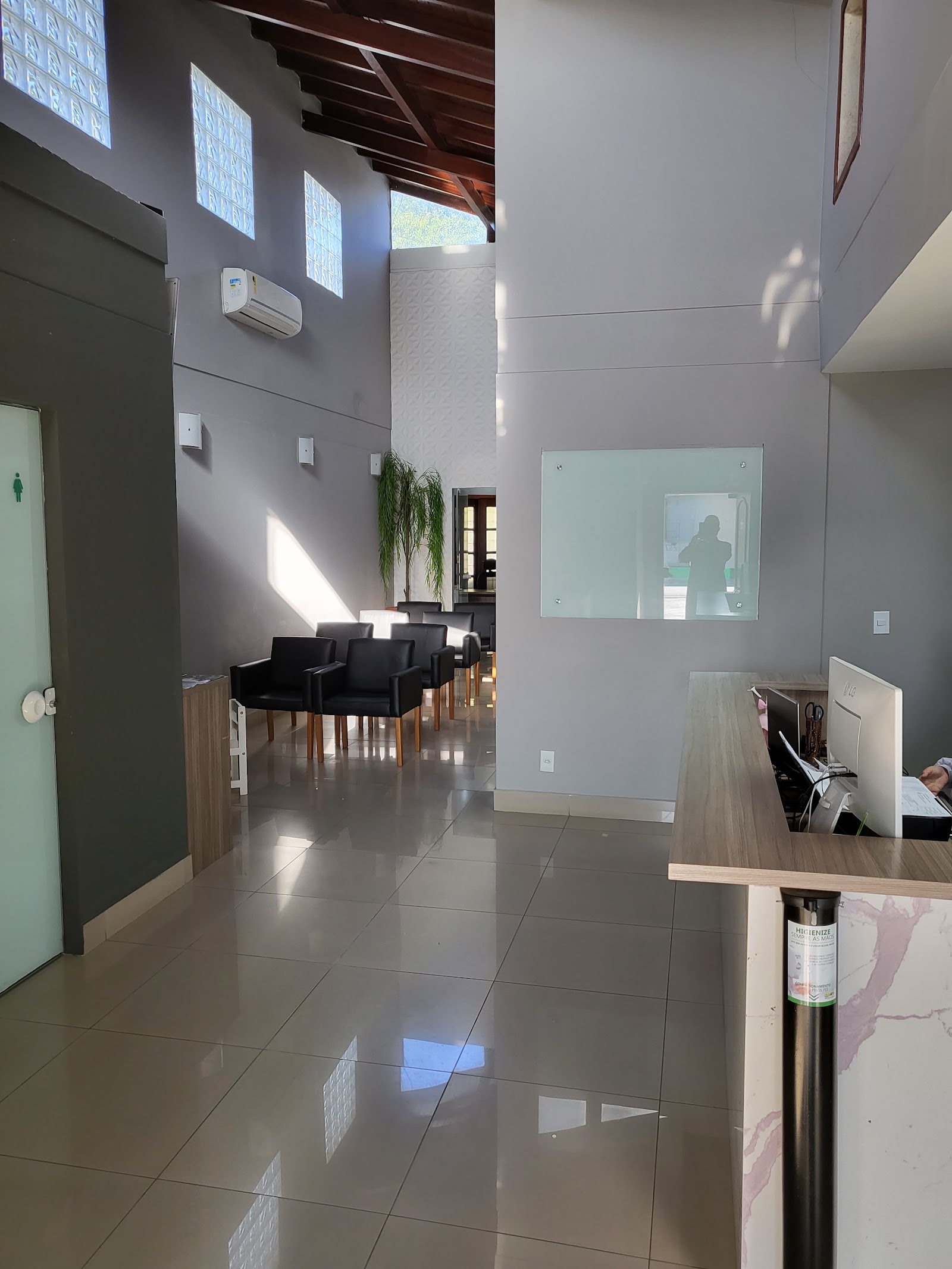 Recepção da clínica com balcão de madeira, sala de espera com poltronas pretas e janelas de vidro no alto